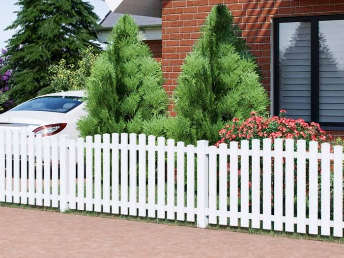 En velplejet have med to koniske stedsegrønne buske og en række røde blomster suppleres af et Stakithegn i Plast "PROFEXline" 180x90/80cm (BUE OP) stakithegn foran. En hvid bil er delvist synlig i venstre side med et murstenshus og et stort vindue i baggrunden.