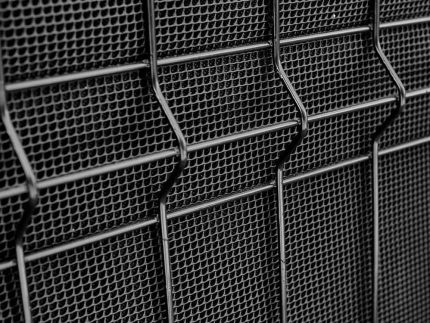 Nærbillede af et industrielt metalnethegn med et gentaget, geometrisk mønster. Metaltrådene danner en række små sekskantede åbninger, hvilket skaber en tekstureret gittereffekt. Billedet er i gråtoner, hvilket understreger de strukturelle detaljer i Støjhegn ECO 18Db Modul.