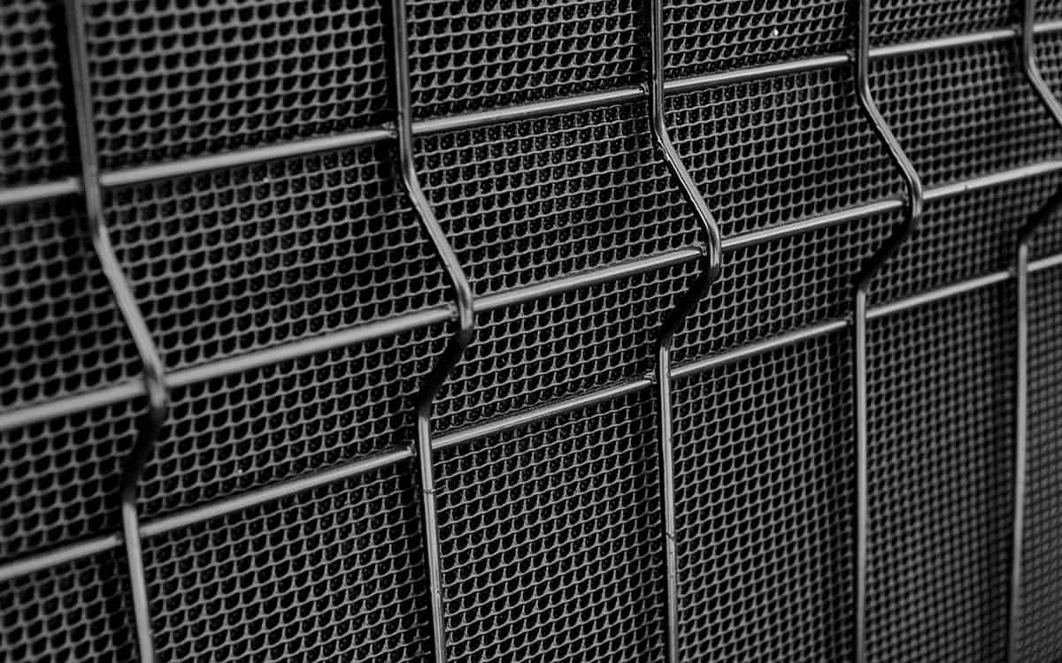 Nærbillede af et industrielt metalnethegn med et gentaget, geometrisk mønster. Metaltrådene danner en række små sekskantede åbninger, hvilket skaber en tekstureret gittereffekt. Billedet er i gråtoner, hvilket understreger de strukturelle detaljer i Støjhegn ECO 18Db Modul.