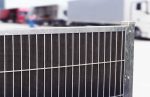 Nærbillede af et metalhegn med netstruktur i et industriområde, potentielt med Støjhegn ECO 18Db Modul. Ude af fokus i baggrunden er et par lastbiler, en rød og en hvid, parkeret ved siden af hinanden. Scenen foreslår en transport- eller logistikindstilling.