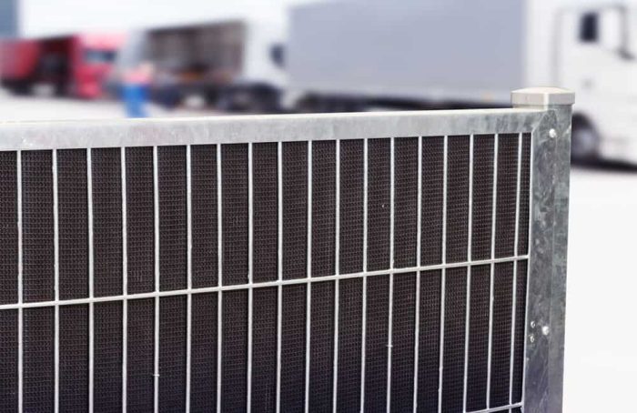Nærbillede af et metalhegn med netstruktur i et industriområde, potentielt med Støjhegn ECO 18Db Modul. Ude af fokus i baggrunden er et par lastbiler, en rød og en hvid, parkeret ved siden af hinanden. Scenen foreslår en transport- eller logistikindstilling.