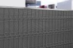 Et nærbillede af et moderne, metallisk mesh-hegn med et geometrisk mønster. Baggrunden viser slørede bygninger og strukturer, der ikke er i fokus, hvilket tilføjer dybde til billedet. Støjhegn ECO 18Db Modul forbedrer både form og funktion.