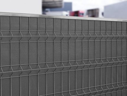 Et nærbillede af et moderne, metallisk mesh-hegn med et geometrisk mønster. Baggrunden viser slørede bygninger og strukturer, der ikke er i fokus, hvilket tilføjer dybde til billedet. Støjhegn ECO 18Db Modul forbedrer både form og funktion.