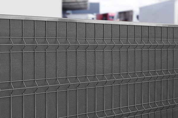 Et nærbillede af et moderne, metallisk mesh-hegn med et geometrisk mønster. Baggrunden viser slørede bygninger og strukturer, der ikke er i fokus, hvilket tilføjer dybde til billedet. Støjhegn ECO 18Db Modul forbedrer både form og funktion.