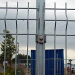 Nærbillede af et 3D Panelhegn 5 mm hegn fastgjort med en bolt og klemme. Bag hegnet ses en blå skibscontainer med et træ til venstre. Himlen er overskyet, og der er nogle tegn i baggrunden nær bunden af hegnet.