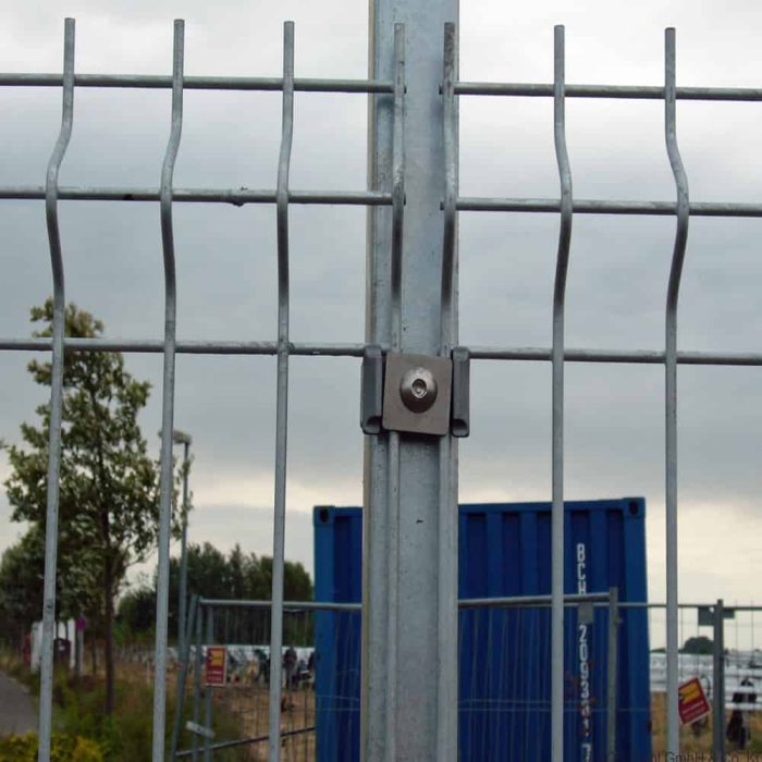 Nærbillede af et 3D Panelhegn 5 mm hegn fastgjort med en bolt og klemme. Bag hegnet ses en blå skibscontainer med et træ til venstre. Himlen er overskyet, og der er nogle tegn i baggrunden nær bunden af hegnet.