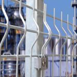 Nærbillede af et 3D Panelhegn 5 mm metaltrådhegn med en robust låsemekanisme på en lodret støttestolpe. Baggrunden er sløret og afslører en del af et industrianlæg med rør og strukturer mod en klar blå himmel.