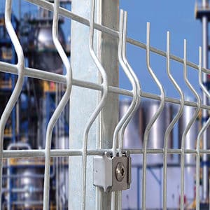 Nærbillede af et 3D Panelhegn 5 mm metaltrådhegn med en robust låsemekanisme på en lodret støttestolpe. Baggrunden er sløret og afslører en del af et industrianlæg med rør og strukturer mod en klar blå himmel.