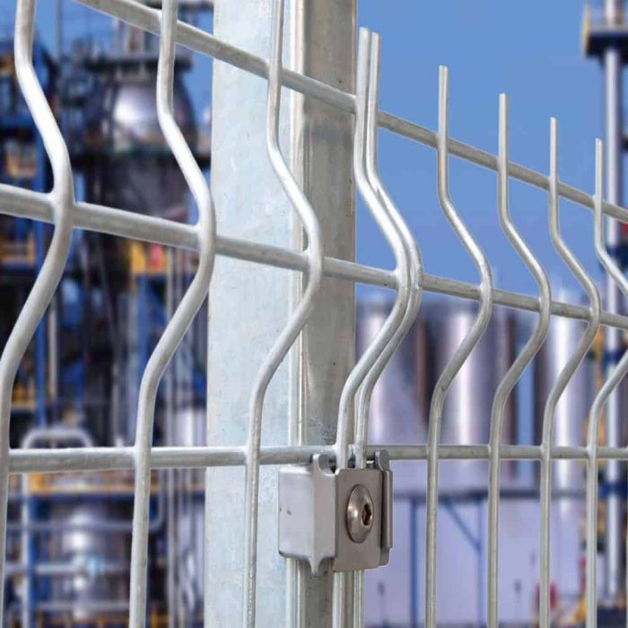 Nærbillede af et 3D Panelhegn 5 mm metaltrådhegn med en robust låsemekanisme på en lodret støttestolpe. Baggrunden er sløret og afslører en del af et industrianlæg med rør og strukturer mod en klar blå himmel.
