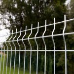 Nærbillede af en sektion af metalhegn med et vekslende mønster af lodrette og bølgede vandrette ledninger. 3D Panelhegn 5 mm, lavet af robuste 5 mm ledninger, skiller sig ud mod de slørede grønne træer og græsset i baggrunden. Billedet fremhæver hegnets struktur og design.