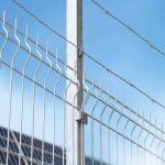 Et nærbillede af et højt 3D Panelhegn 5 mm hegn, med 5 mm lodrette trådstænger og pigtråd, der løber hen over toppen, sat mod en klar blå himmel. Denne struktur er forankret af en robust metallisk stolpe.