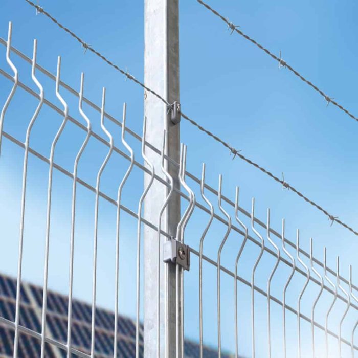 Et nærbillede af et højt 3D Panelhegn 5 mm hegn, med 5 mm lodrette trådstænger og pigtråd, der løber hen over toppen, sat mod en klar blå himmel. Denne struktur er forankret af en robust metallisk stolpe.