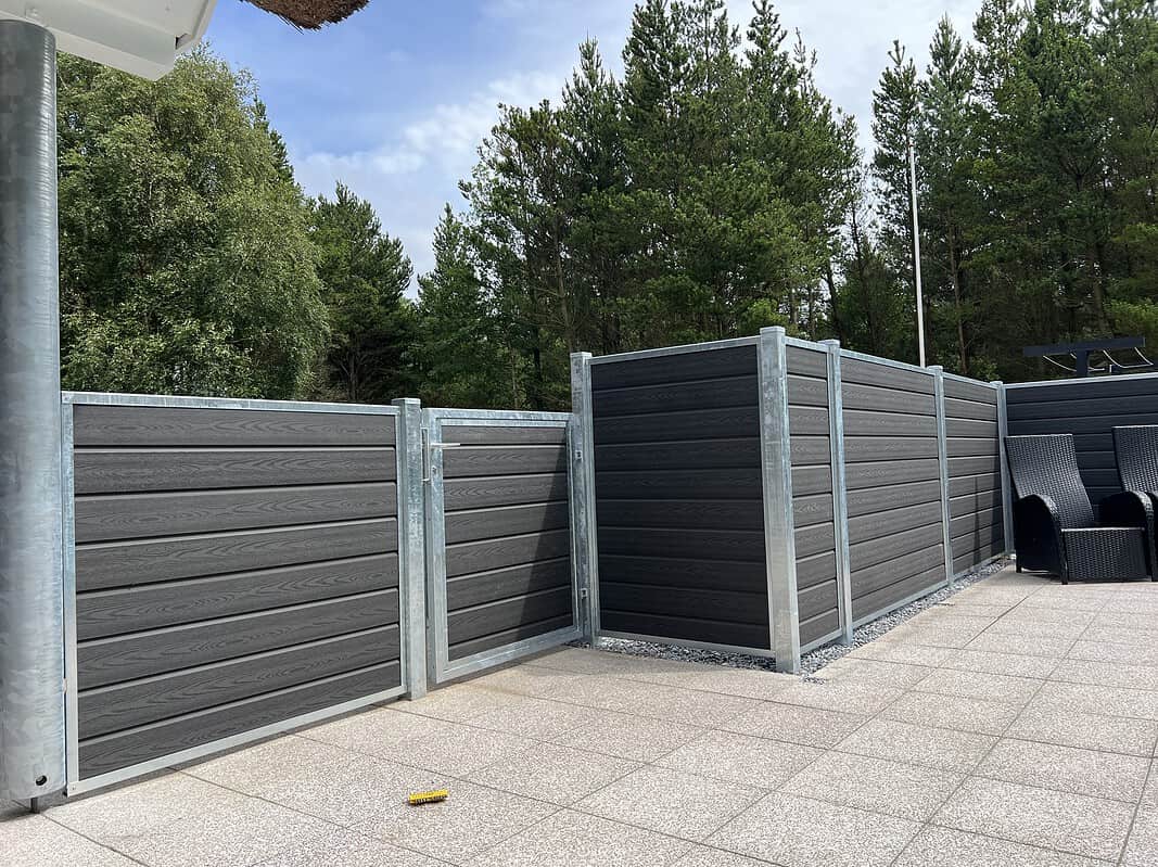 Et moderne udendørs område har mørkegrå komposithegn med metalrammer, suppleret med "Låge Varmgalvaniseret Type 1 - Inkl. Stolper og Tilbehør - 25mm Komposit Brædder." Hegnet omslutter en del af en flisebelagt terrasse. I baggrunden fuldender høje grønne træer og en blå himmel med nogle skyer de fredfyldte omgivelser. To sorte udendørs stole er tæt på hegnet.