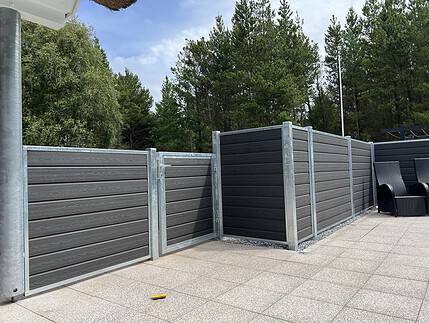 Et moderne udendørs område har mørkegrå komposithegn med metalrammer, suppleret med "Låge Varmgalvaniseret Type 1 - Inkl. Stolper og Tilbehør - 25mm Komposit Brædder." Hegnet omslutter en del af en flisebelagt terrasse. I baggrunden fuldender høje grønne træer og en blå himmel med nogle skyer de fredfyldte omgivelser. To sorte udendørs stole er tæt på hegnet.