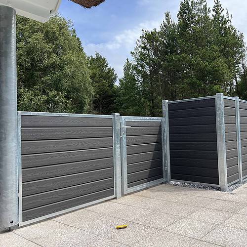 Et moderne udendørs område har mørkegrå komposithegn med metalrammer, suppleret med "Låge Varmgalvaniseret Type 1 - Inkl. Stolper og Tilbehør - 25mm Komposit Brædder." Hegnet omslutter en del af en flisebelagt terrasse. I baggrunden fuldender høje grønne træer og en blå himmel med nogle skyer de fredfyldte omgivelser. To sorte udendørs stole er tæt på hegnet.