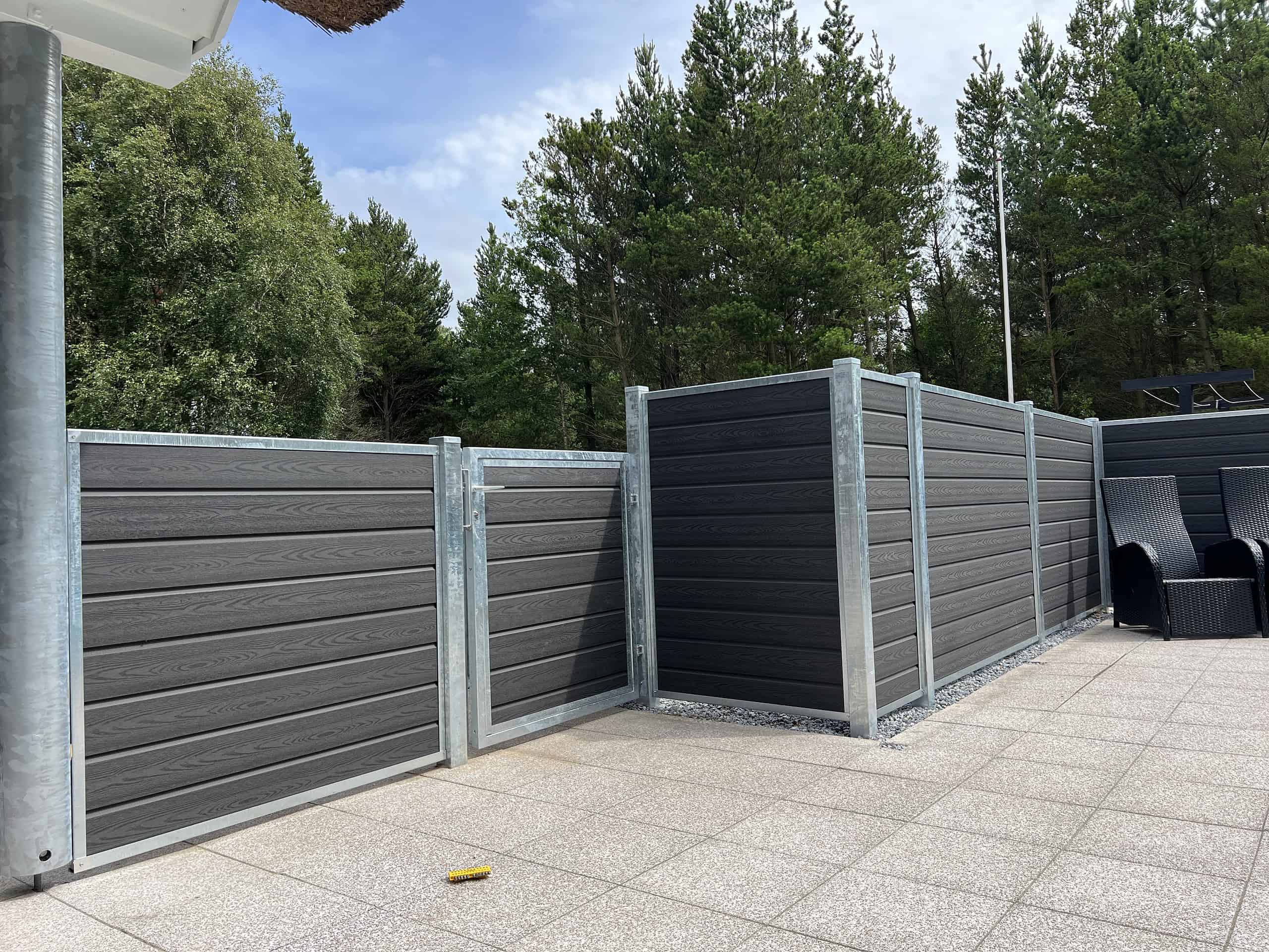 Et moderne udendørs område har mørkegrå komposithegn med metalrammer, suppleret med "Låge Varmgalvaniseret Type 1 - Inkl. Stolper og Tilbehør - 25mm Komposit Brædder." Hegnet omslutter en del af en flisebelagt terrasse. I baggrunden fuldender høje grønne træer og en blå himmel med nogle skyer de fredfyldte omgivelser. To sorte udendørs stole er tæt på hegnet.