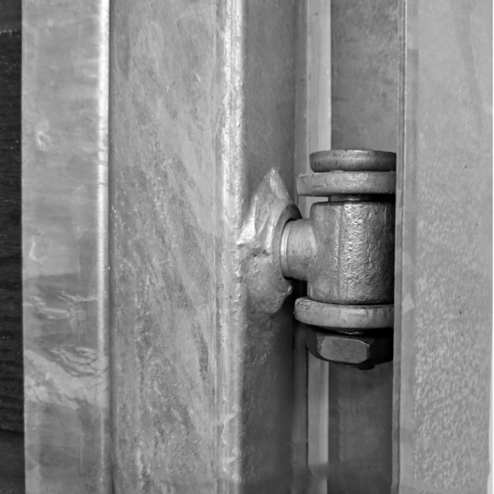 Nærbillede af et kraftigt metalhængsel fastgjort til to store metalflader, potentielt en Lågeramme med lås - Varmgalvaniseret HDG - til 25mm komposit brædder. Hængslet er sikret med bolte og ser ud til at være en del af en strukturel eller industriel installation. Det sort-hvide billede fremhæver tekstur og slid på metaloverfladerne.