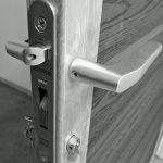 Et nærbillede af en sort-hvid dørlåsemekanisme. Det metalliske dørhåndtag og låsemekanismen er afbildet på kanten af en åben trædør. Mærket "LOCINOX" og "100% INOX" er indgraveret på låsepladen, designet til at passe perfekt til Lågeramme med lås - Varmgalvaniseret HDG - til 25mm komposit brædder.