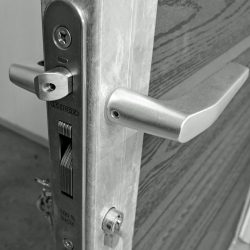 Et nærbillede af en sort-hvid dørlåsemekanisme. Det metalliske dørhåndtag og låsemekanismen er afbildet på kanten af en åben trædør. Mærket "LOCINOX" og "100% INOX" er indgraveret på låsepladen, designet til at passe perfekt til Lågeramme med lås - Varmgalvaniseret HDG - til 25mm komposit brædder.