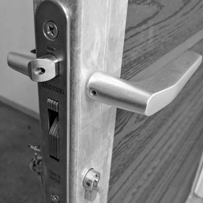 Et nærbillede af en sort-hvid dørlåsemekanisme. Det metalliske dørhåndtag og låsemekanismen er afbildet på kanten af en åben trædør. Mærket "LOCINOX" og "100% INOX" er indgraveret på låsepladen, designet til at passe perfekt til Lågeramme med lås - Varmgalvaniseret HDG - til 25mm komposit brædder.