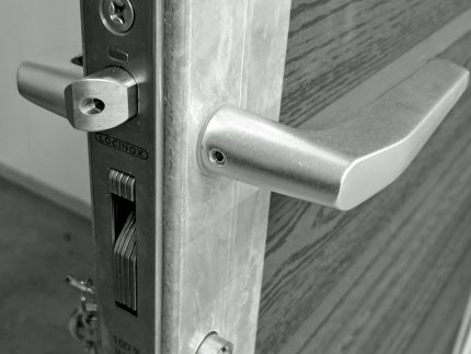 Et nærbillede af en sort-hvid dørlåsemekanisme. Det metalliske dørhåndtag og låsemekanismen er afbildet på kanten af en åben trædør. Mærket "LOCINOX" og "100% INOX" er indgraveret på låsepladen, designet til at passe perfekt til Lågeramme med lås - Varmgalvaniseret HDG - til 25mm komposit brædder.