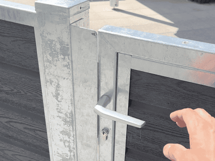 Et nærbillede af Låge Varmgalvaniseret Type 1 - Inkl. Stolper og Tilbehør - 25mm Komposit Brædder med håndtag, set fra en let forhøjet vinkel. En hånd rækker ud mod håndtaget, klar til at åbne lågen. Baggrunden omfatter en flisebelagt grund og nogle udendørsmøbler, hvilket indikerer en udendørs indstilling.