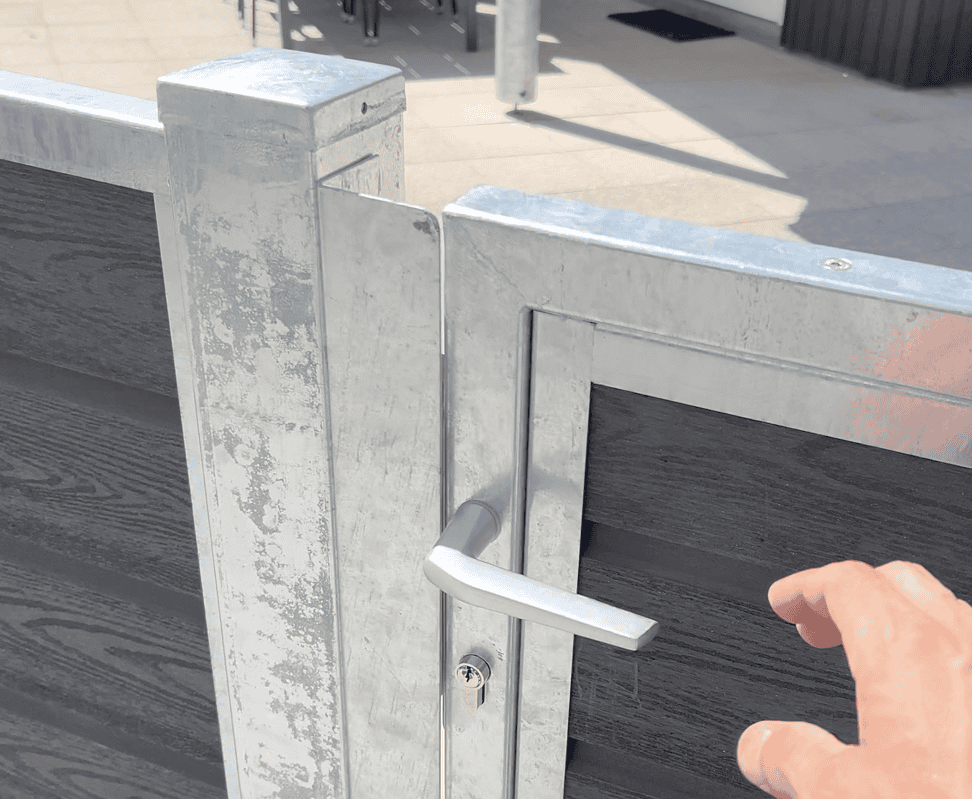 Et nærbillede af Låge Varmgalvaniseret Type 1 - Inkl. Stolper og Tilbehør - 25mm Komposit Brædder med håndtag, set fra en let forhøjet vinkel. En hånd rækker ud mod håndtaget, klar til at åbne lågen. Baggrunden omfatter en flisebelagt grund og nogle udendørsmøbler, hvilket indikerer en udendørs indstilling.