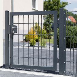 En moderne, sort metalport med lodrette og vandrette stænger er en del af et hegn, der omslutter en anlagt gård. Bag porten er grønne buske, små træer og et glimt af et boligområde. Porten er fastgjort til en beige væg på den ene side, hvilket eksemplificerer kvalitet har hegn og låger i sit design.