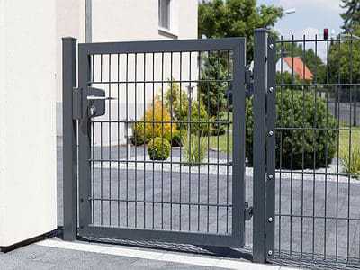 En moderne, sort metalport med lodrette og vandrette stænger er en del af et hegn, der omslutter en anlagt gård. Bag porten er grønne buske, små træer og et glimt af et boligområde. Porten er fastgjort til en beige væg på den ene side, hvilket eksemplificerer kvalitet har hegn og låger i sit design.