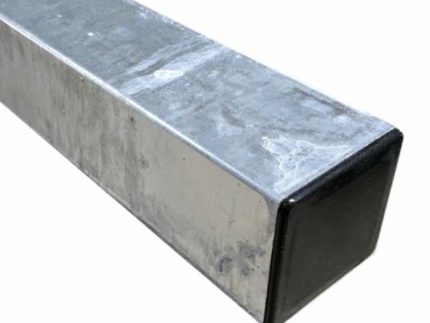 Nærbillede af et firkantet metalrør, stålstolpe varmgalv. 100x100x2600mm, med en skinnende, reflekterende overflade. Røret er hult og har en sort plastikhætte i den ene ende. Metaloverfladen fremstår let slidt med enkelte synlige ridser og mærker.