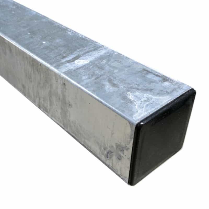 Nærbillede af et firkantet metalrør, stålstolpe varmgalv. 100x100x2600mm, med en skinnende, reflekterende overflade. Røret er hult og har en sort plastikhætte i den ene ende. Metaloverfladen fremstår let slidt med enkelte synlige ridser og mærker.