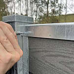 Ved hjælp af U-skinne top/bund - varmgalvaniseret fastgør en person et top/bund skinne på et komposit hegn med præcision og integrerer det problemfrit. Træer og en bygning er sløret i baggrunden.