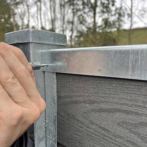 Ved hjælp af U-skinne top/bund - varmgalvaniseret fastgør en person et top/bund skinne på et komposit hegn med præcision og integrerer det problemfrit. Træer og en bygning er sløret i baggrunden.