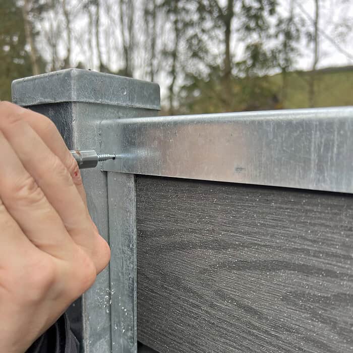 Ved hjælp af U-skinne top/bund - varmgalvaniseret fastgør en person et top/bund skinne på et komposit hegn med præcision og integrerer det problemfrit. Træer og en bygning er sløret i baggrunden.