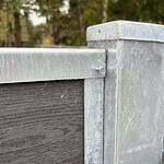 Et nærbillede af en U-skinne top/bund - varmgalvaniseret metal og mørkt træ hegnshjørne med en bolt. De galvaniserede sektioner udviser let slid og edderkoppespind, mens blødt slørede træer i baggrunden antyder udendørs omgivelser.