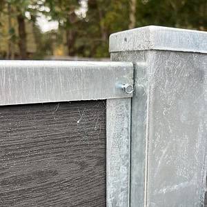 Et nærbillede af en U-skinne top/bund - varmgalvaniseret metal og mørkt træ hegnshjørne med en bolt. De galvaniserede sektioner udviser let slid og edderkoppespind, mens blødt slørede træer i baggrunden antyder udendørs omgivelser.