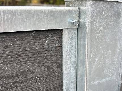 Et nærbillede af en U-skinne top/bund - varmgalvaniseret metal og mørkt træ hegnshjørne med en bolt. De galvaniserede sektioner udviser let slid og edderkoppespind, mens blødt slørede træer i baggrunden antyder udendørs omgivelser.