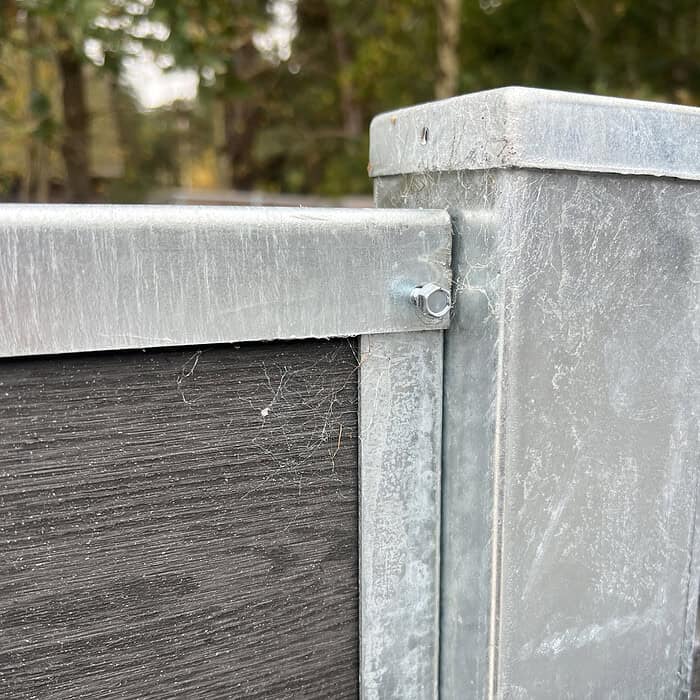 Et nærbillede af en U-skinne top/bund - varmgalvaniseret metal og mørkt træ hegnshjørne med en bolt. De galvaniserede sektioner udviser let slid og edderkoppespind, mens blødt slørede træer i baggrunden antyder udendørs omgivelser.
