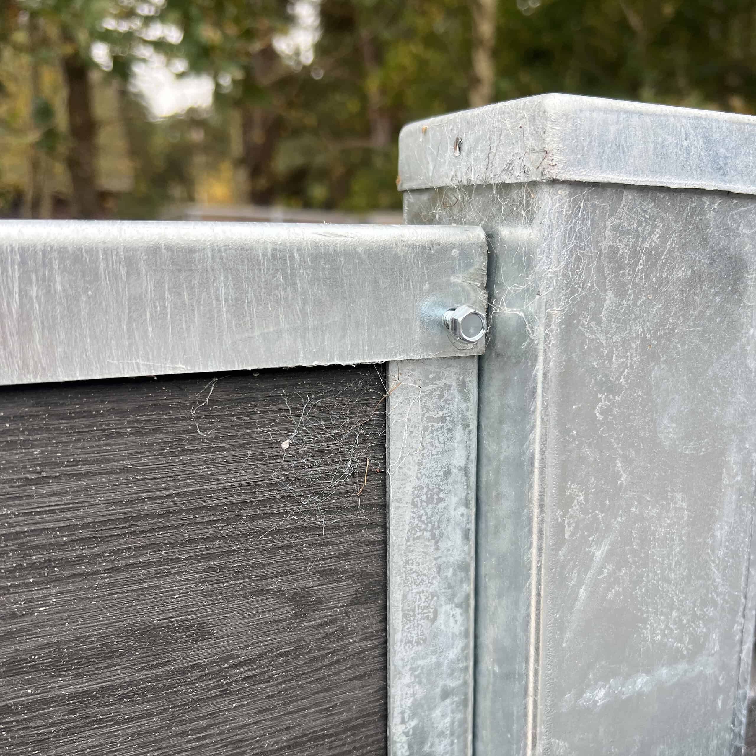 Et nærbillede af en U-skinne top/bund - varmgalvaniseret metal og mørkt træ hegnshjørne med en bolt. De galvaniserede sektioner udviser let slid og edderkoppespind, mens blødt slørede træer i baggrunden antyder udendørs omgivelser.