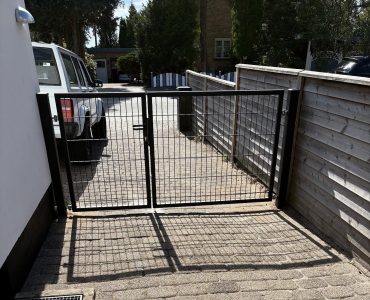 En sort metalport blokerer indgangen til en indkørsel med brosten. En hvid bil er parkeret til venstre, og træhegn løber langs begge sider og fører mod et boligområde med træer og huse.