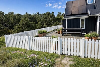 Et sort hus med solpaneler ligger ved et hvidt stakit, omgivet af potteplanter og grønne planter. En terrasse med bord og bænke er synlig, og træer fylder baggrunden under en delvist overskyet himmel.