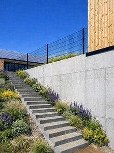 Betontrapper omgivet af blomstrende planter og græs går op langs en høj betonmur med et sort trådhegn, som fører til moderne bygninger under en klar blå himmel.