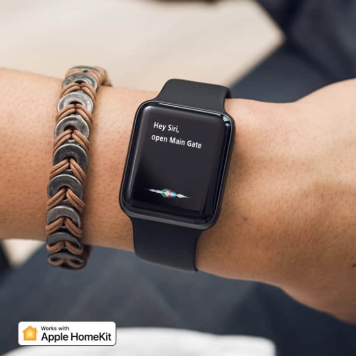 En person, der bærer et sort smartwatch og et flettet armbånd, vises. Urskærmen viser meddelelsen "Hey Siri, åben hovedporten." Nederst til venstre er der et "Works with Apple HomeKit"-logo. Denne opsætning integreres problemfrit med ismartgate LITE - Fjernstyret Låge & portåbner via. Din Smartphone eller PC - (Max 1 tilslutning af låge eller port) til dine fjernbetjente port- og garageportbehov.
