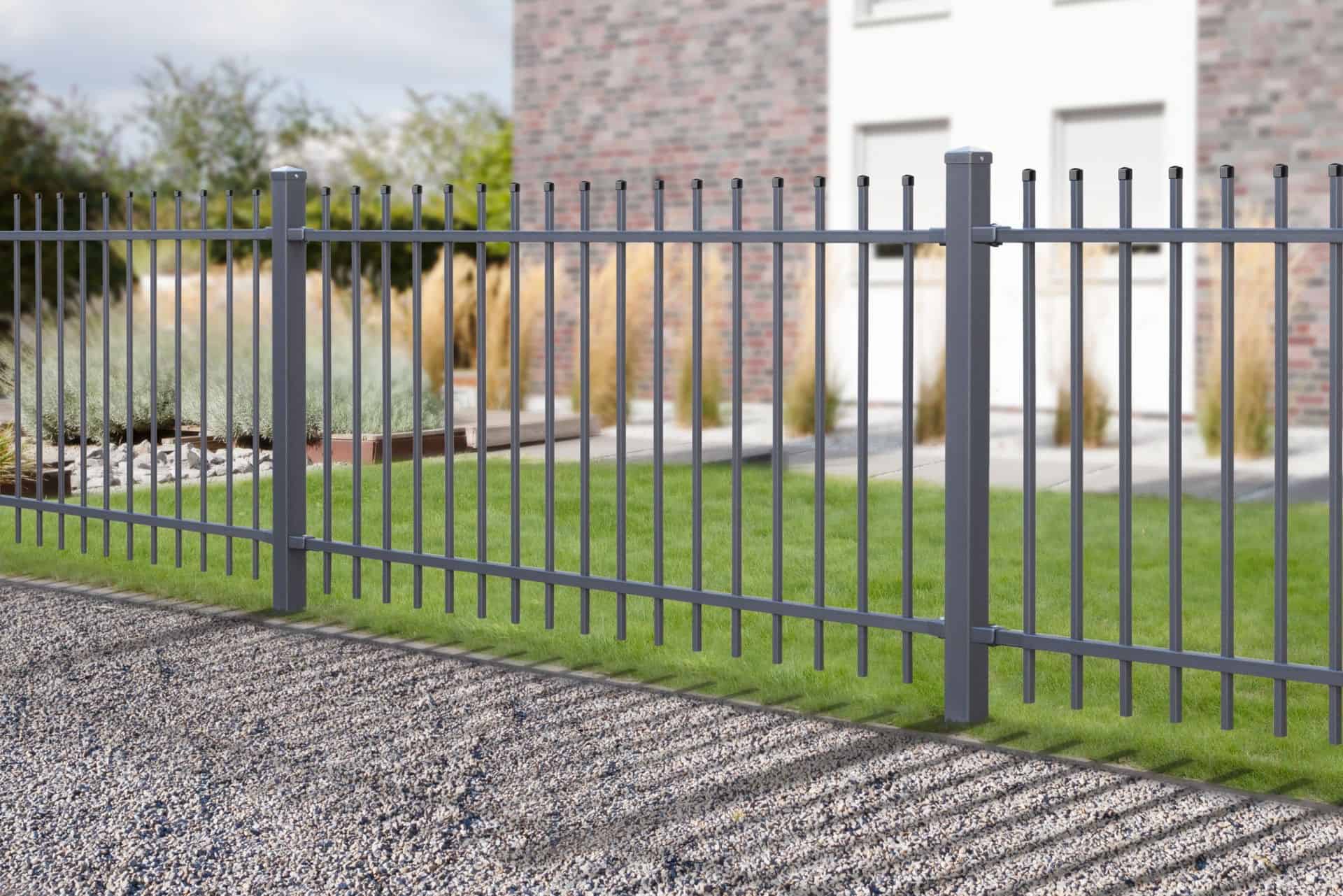 En sort smedejernshegn med lodrette stænger omslutter en grøn, pænt trimmet græsplæne. Hegnet skygger på en grussti foran, og i baggrunden ses en moderne murstensbygning med vinduer. Nogle dekorative planter optræder også i baggrunden, hvilket skaber en indbydende forside.