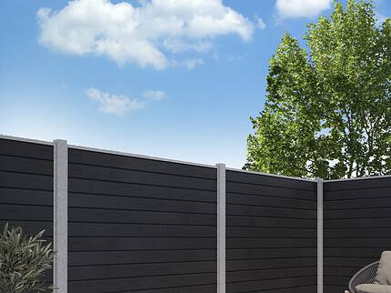 En moderne baggård har et sort vandret lamelhegn, konstrueret som Komposit Hegn med galvaniseret stålstolper & u-skinner - Type 1, understøttet af grå stolper. Grunden er belagt med grå stenfliser. Et træ med grønne blade rejser sig over hegnet til højre, og himlen er lysende blå med spredte hvide skyer.