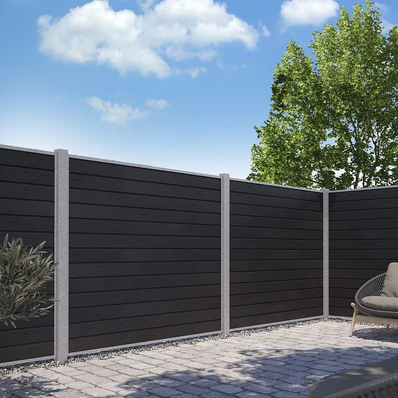 En moderne baggård har et sort vandret lamelhegn, konstrueret som Komposit Hegn med galvaniseret stålstolper & u-skinner - Type 1, understøttet af grå stolper. Grunden er belagt med grå stenfliser. Et træ med grønne blade rejser sig over hegnet til højre, og himlen er lysende blå med spredte hvide skyer.