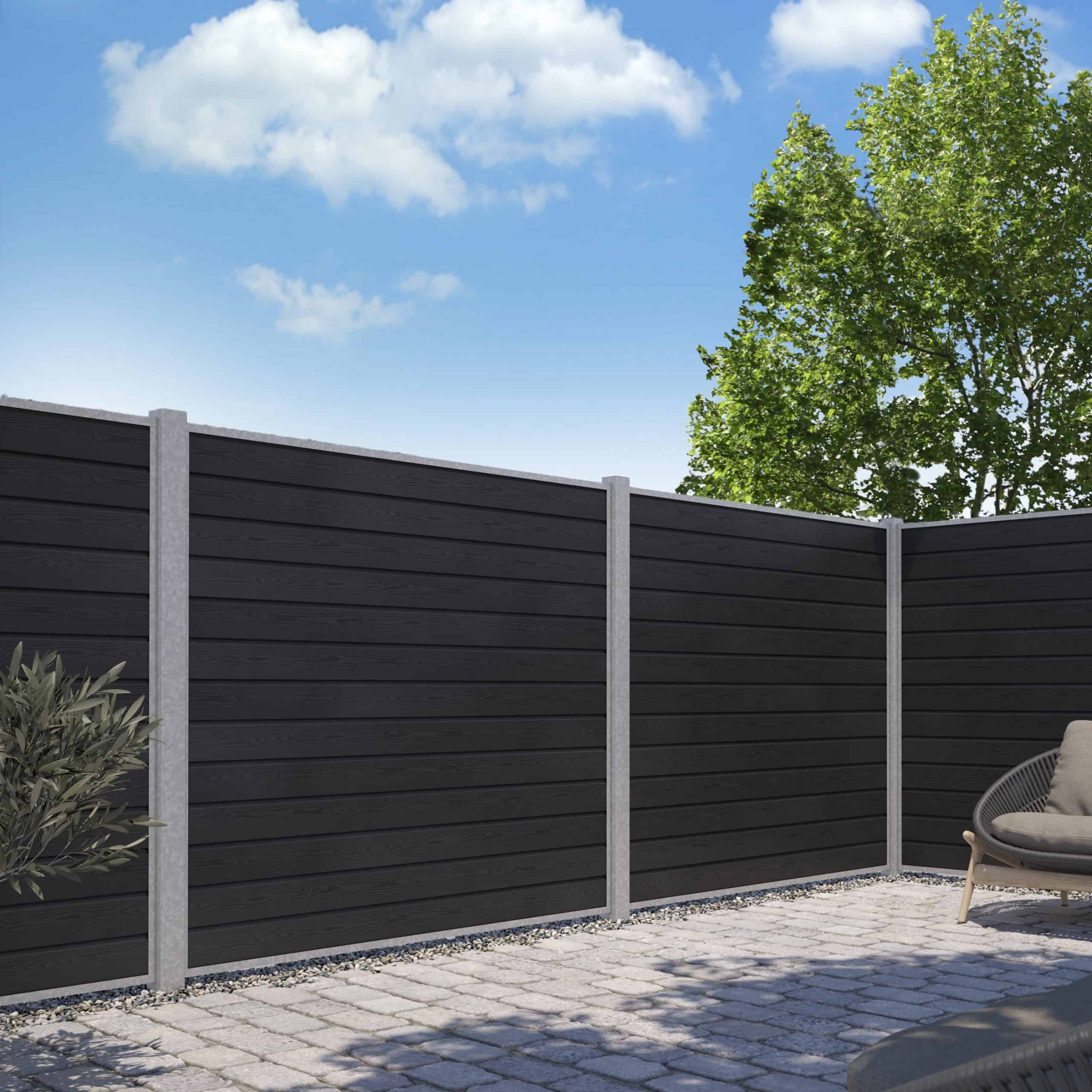 En moderne baggård har et sort vandret lamelhegn, konstrueret som Komposit Hegn med galvaniseret stålstolper & u-skinner - Type 1, understøttet af grå stolper. Grunden er belagt med grå stenfliser. Et træ med grønne blade rejser sig over hegnet til højre, og himlen er lysende blå med spredte hvide skyer.