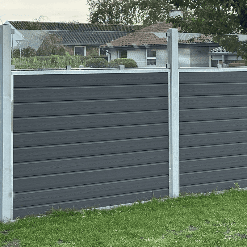 Nærbillede af en mørkegrå, moderne Komposit Hegn med klartglas - Inkl. galvaniseret stålstolper u-skind med vandrette paneler. Hegnet er sat på en grøn græsplæne med huse og træer synlige i baggrunden.