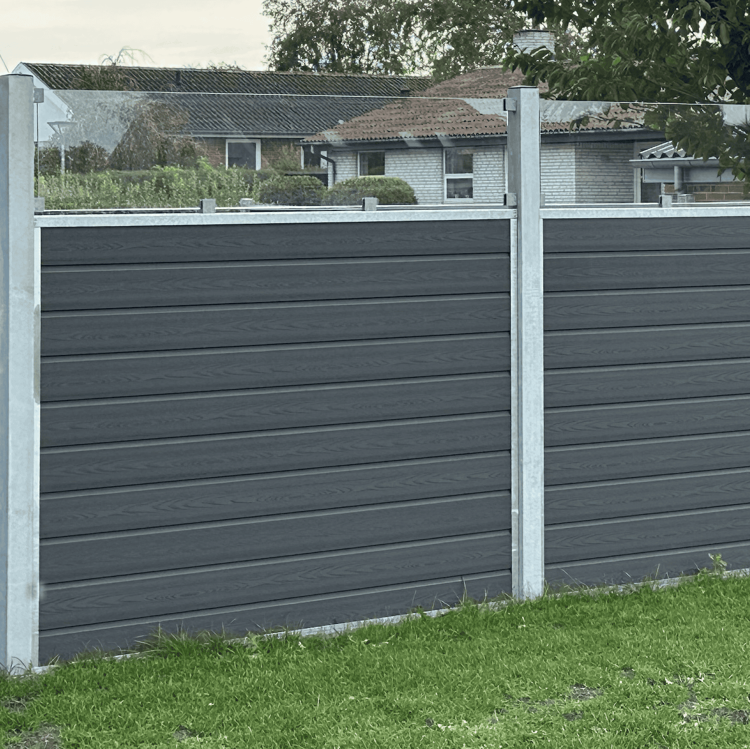 Nærbillede af en mørkegrå, moderne Komposit Hegn med klartglas - Inkl. galvaniseret stålstolper u-skind med vandrette paneler. Hegnet er sat på en grøn græsplæne med huse og træer synlige i baggrunden.