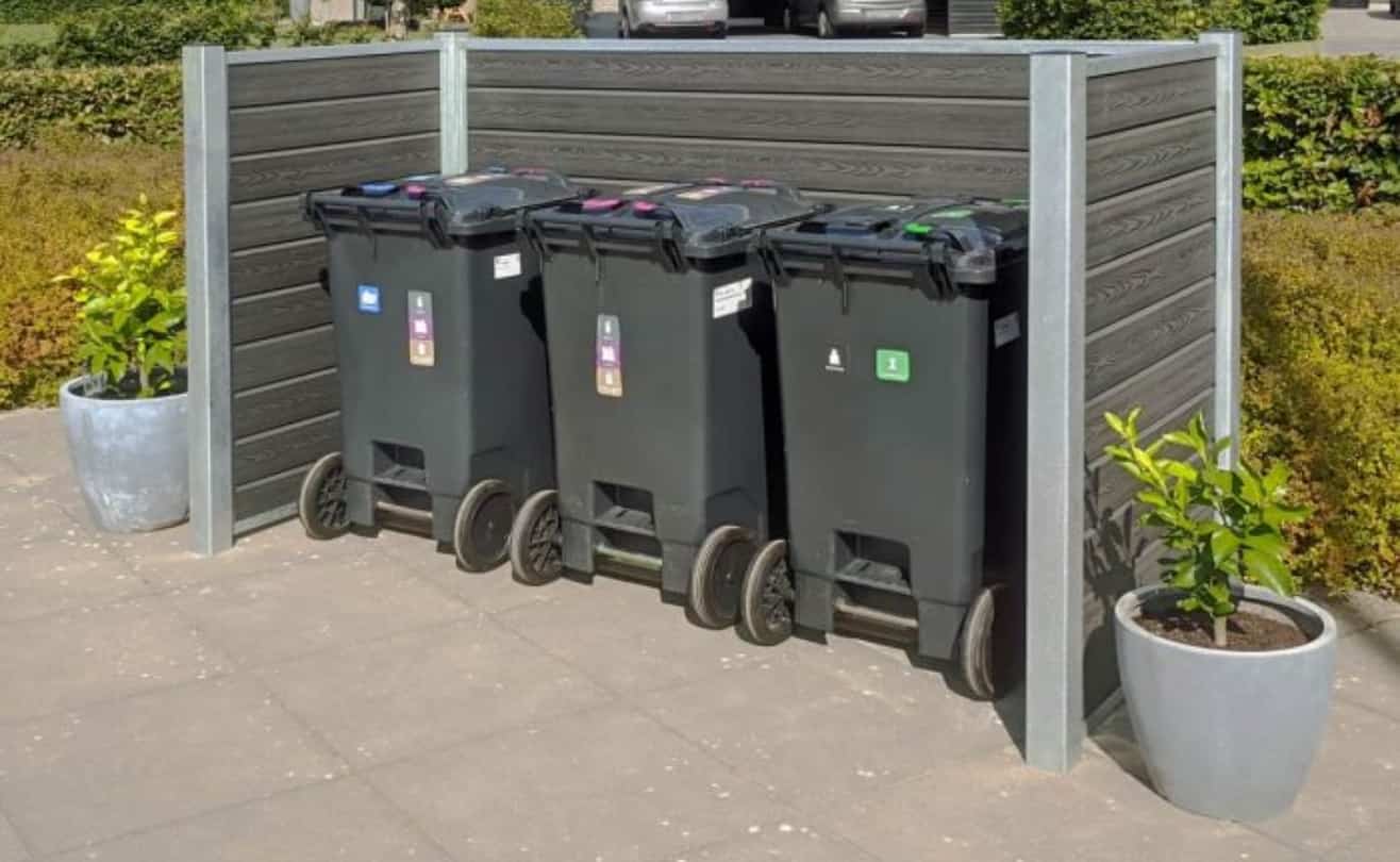 Tre store skraldespande på hjul står side om side ved siden af et hegnspanel. De er sorte og har farvede låg og klistermærker, der angiver forskellige typer affald. To store potteplanter er placeret i hver ende af beholderne, hvilket tilføjer grønt til scenen. Fordele ved at benytte skraldespandsskjulere omfatter forbedret æstetik og organisation.